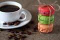 Macaron three pieces red, green and orange tied with rope and mug with coffee, scattered beans and saucer on burlap tablecloth Royalty Free Stock Photo