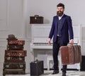 Macho elegant on smiling face stands near pile of vintage suitcase, holds suitcase. Man, traveller with beard and