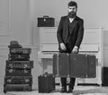 Macho elegant on strict face stands near pile of vintage suitcase, holds suitcase. Man, traveller with beard and