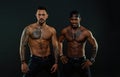 Machos with muscular tattooed torsos look attractive, dark background. Athletes on confident faces with nude muscular