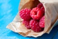 Macro photo of paper cornet with many freshly picked raw sweet raspberries on blue painted wood textured