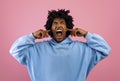 Mad black teenager shouting, covering ears and closing eyes over pink studio background