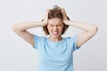 Mad crazy young woman in blue t shirt with hands on head and eyes closed standing and suffering from headache isolated over white Royalty Free Stock Photo