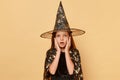 Magical wizard and sorcerer. Astonished little girl wearing witch costume and carnival cone hat isolated over beige background Royalty Free Stock Photo