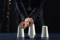 Magician showing tricks with cups on dark background