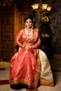 Magnificent young Indian bride in luxurious dress and precious jewellery is sitting in a chair in a luxury apartment. Classic