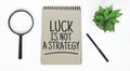 A magnifying glass, a blank brown notebook on a white table. Luck is not A Strategy sign