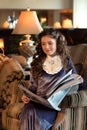 Maiden student in a retro dress with a jabot sits in an antique chair and reads a newspaper pensively dreams