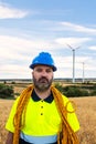 Maintenance worker standing in wind turbine farm field with climbing equipment: helmet and rope. Looking at camera.