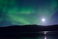 Majestic aurora borealis dancing beside full moon over mountain and calm fjord