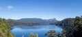 Majestic nature view of a huge clear blue lake reflecting the forest that surrounds it and the mountain range in the distance, in Royalty Free Stock Photo