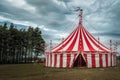 A majestic red and white circus tent stands against a backdrop of tall pine trees and a cloudy sky. Royalty Free Stock Photo