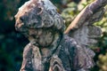 Majestic view of a weathered statue of a small angel against a background of dark foliage. Dramatic unusual scene. Beauty statue