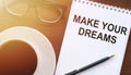 Make your dreams, the text is written on a notebook with glasses and coffee on the table. Sunlight is falling. Concept of desire