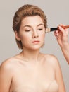 Makeup artist applies foundation on the skin of a girl with short blonde hair. Shot in a studio on a light background