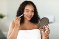 Daily makeup. Beautiful oversize black lady applying blush with makeup brush, holding mirror and looking at camera Royalty Free Stock Photo