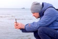 Man blogger in winter clothes at sea beach shooting video of waves on phone.