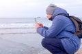 Man blogger in winter clothes at sea beach shooting video of waves on phone.