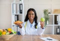 Making healthy food choices. Beautiful female nutritionist holding croissant and apple at table in medical office Royalty Free Stock Photo