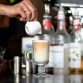 Making latte coffee. Barista pours freshly brewed coffee into whipped milk froth in front of bottles on blurred Royalty Free Stock Photo