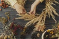 Making rustic autumn boho wreath flat lay. Hands holding dry grass and making stylish wreath with wildflowers, herbs and berries