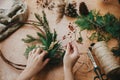 Making rustic christmas wreath. Hands holding berries, fir branches, pine cones, thread, scissors on wooden table. Authentic