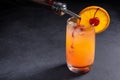 Making Tequila Sunrise Cocktail. Grenadine slowly poured into a glass of ice, orange juice and tequila. Dark stone background with Royalty Free Stock Photo