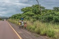 View of woman with children walks with head load, along roadside, tropical landscape as background