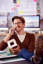 Male Architect In Office With Feet On Desk Taking Call On Mobile Phone And Looking At Wooden Model