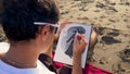 Male artist sitting on a sand beach painting chracter with watercolor and paintbrush. Game developer 2D artist making up