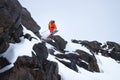 Male athlete skier in an orange trigger with a gray backpack and ski poles jumping from a high rocky snowy slope against Royalty Free Stock Photo