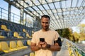 Male athlete uses app on phone after fitness class at stadium, trainer in headphones uses smartphone to listen to music