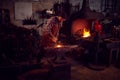 Male Blacksmith Hammering Metalwork On Anvil With Blazing Forge In Background