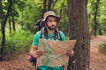 Male brunet bearded confused tourist got lost in the forest, holding map, looking far, trying to find the way. He has a backpack,