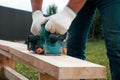 Male carpenter electric planers planed board on the lawn near the house. Close-up view of the tool and flying sawdust