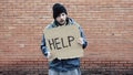 Homeless holds piece of cardboard with inscription Help. Refugee is at brick wall looking for job. Male tramp in dirty clothes,