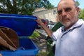 Male Caucasian senior citizen with a frown holding open the lid of a blue recycling trash can with cardboard inside