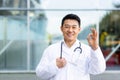 male cheerful asian doctor smiling and looking at camera showing ok hand sign on modern clinic outdoor background Royalty Free Stock Photo