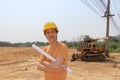 Male civil engineer wear the yellow helmet and cross one arm with project drafts while in hand standing on background of front