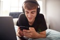 Male College Student Wearing Headphones Lies On Bed In Shared House With Mobile Phone Using Laptop Royalty Free Stock Photo