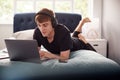 Male College Student Wearing Headphones Lies On Bed In Shared House Working On Laptop Royalty Free Stock Photo