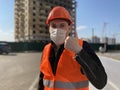 Male construction worker in overalls and in medical mask showing thumbs up on background of house under construction Royalty Free Stock Photo