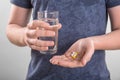 A male in a dark blue T-shirt is holding several pills and capsules, and a big glass of water on a gray background. Royalty Free Stock Photo