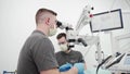 Male dentist with assistant operating patient tooth using microscope at the modern dental clinic. Doctor cleans the