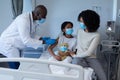 Male doctor giving covid vaccination to sick girl in hospital bed with mother, all in face masks Royalty Free Stock Photo