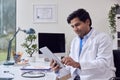 Male Doctor Or GP Wearing White Coat Sitting At Desk In Office Using Digital Tablet Royalty Free Stock Photo