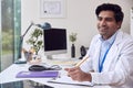 Male Doctor Or GP Wearing White Coat Sitting At Desk In Office Writing Notes On Clipboard Royalty Free Stock Photo