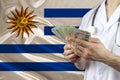 Male doctor holding a fan of dollar banknotes in his hands against the background of the silk national flag of Uruguay, concept of