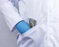 Male doctor in a white medical coat and blue latex gloves puts a stack of US dollars in his coat pocket Royalty Free Stock Photo