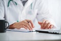 Male doctor working with laptop behind desk, writing on paperwork in a hospital.